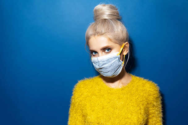 Studio portrait of young blonde girl with hair bun wearing medical respiratory safety mask on face against coronavirus. Background of phantom blue color. - 写真・画像