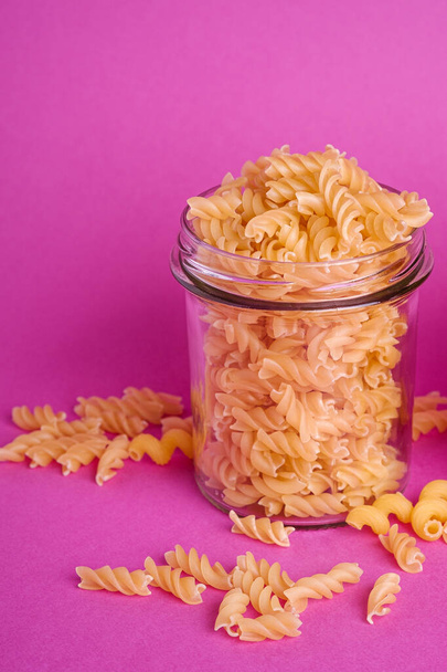 One glass jar with fusilli uncooked golden wheat pasta on minimal pink background, angle view macro - Photo, Image