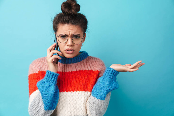 Image of unhappy beautiful woman in eyeglasses talking on smartphone isolated over blue background - Fotografie, Obrázek