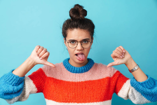 Image of displeased beautiful woman in eyeglasses showing her tongue and thumbs down isolated over blue background - 写真・画像