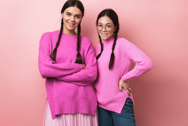 Image of two happy teenage girls with braids in casual clothes smiling at camera isolated over pink background - Photo, image