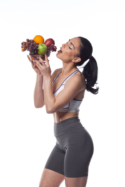 Sexy brunette woman in fitness suit eating fruits. Studio shoot isolated on white background. - Valokuva, kuva