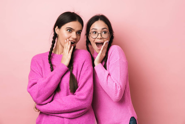 Image of two happy teenage girls with braids expressing wonder while looking at copyspace isolated over pink background - Foto, Imagem