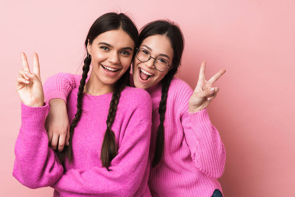 Image of two happy teenage girls with braids in casual clothes smiling and gesturing peace fingers isolated over pink background - Photo, Image