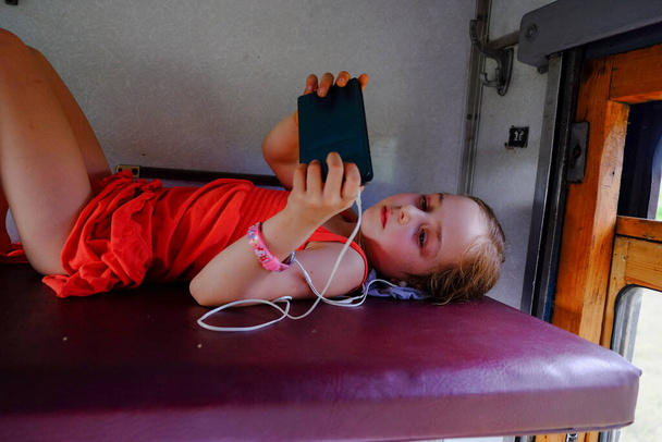 Roztomilá holka ležící na horní polici ve vlaku. Letní dovolená a pojetí cestování. dívka ve vlaku leží na horní polici. Dívka ve věku 5 nebo 6 let jezdí vlakem. Dospívající cestují. Smartphone - Fotografie, Obrázek