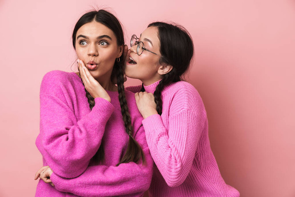 Two cheerful cute teenage girls standing isolated over pink background, telling secrets - Фото, изображение