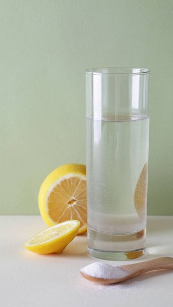 Vitamina C en polvo sobre cuchara de madera con vaso de agua y limón cortado sobre fondo verde y blanco
 - Foto, Imagen