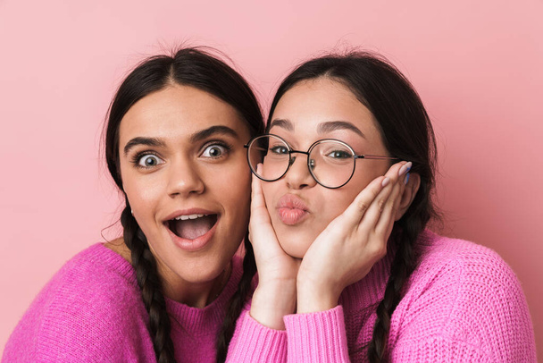 Image of two happy teenage girls with braids in casual clothes smiling at camera isolated over pink background - Zdjęcie, obraz