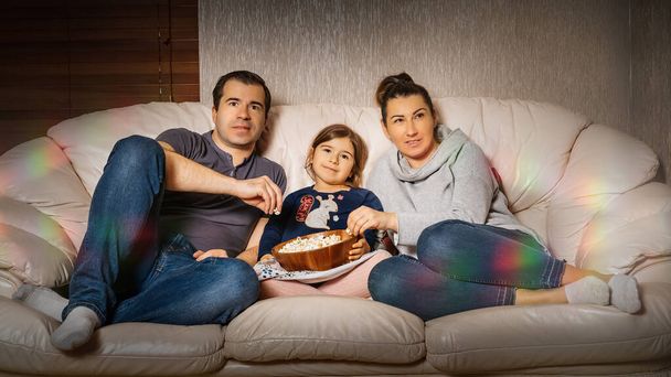 家族はソファの上に一緒に座って、ポップコーンを食べて、彼らの家の快適さから映画を見ています。 - 写真・画像