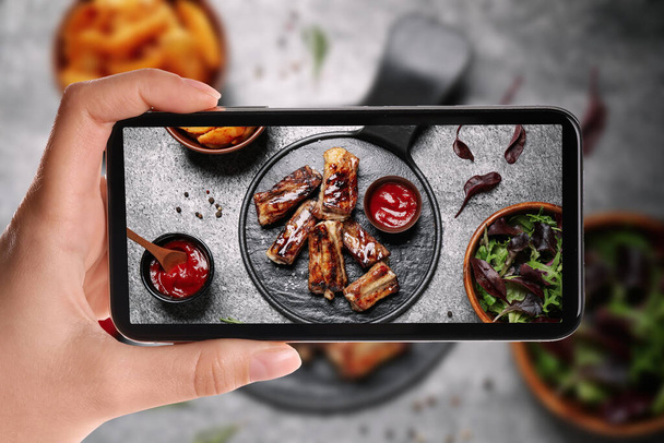 Blogger taking picture of delicious grilled ribs at table, closeup. Food photography - Фото, изображение