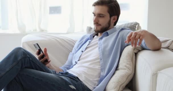 Happy millennial guy relaxing on sofa holding using smart phone - Metraje, vídeo
