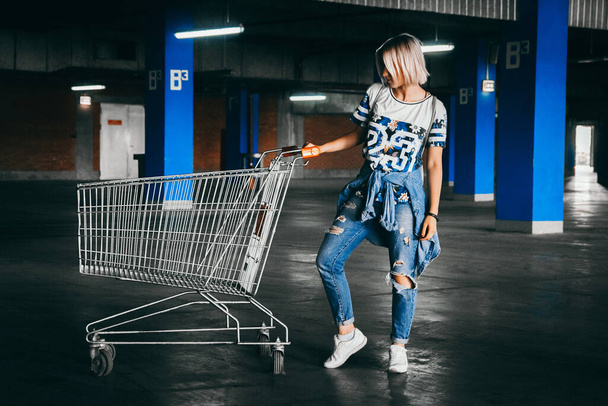 Mooi meisje met kort wit haar gekleed in jeans met winkelwagentjes op de parkeerplaats. Positief jong meisje in de supermarkt trolley. - Foto, afbeelding