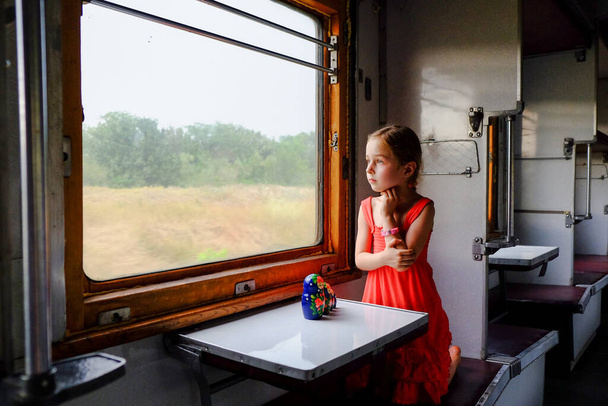 Cute girl in the train. Summer Vacation and Travel Concept. A girl of 5 or 6 years old rides on a train. Teen travels. Little girl with long hair. Child portrait - Photo, Image
