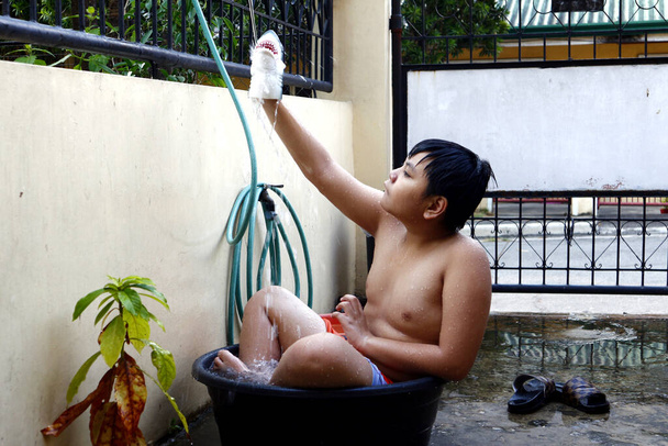 Foto de um jovem menino asiático esfriando em uma bacia de água e uma mangueira de água como um chuveiro improvisado para bater o calor do verão
. - Foto, Imagem