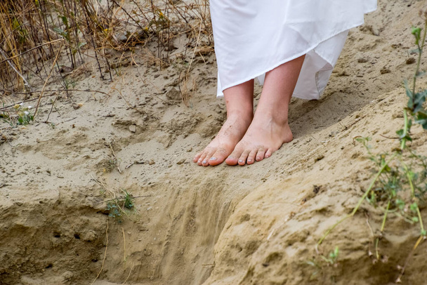 Pernas de uma menina em um vestido branco na borda de um penhasco. A areia está caindo da borda do penhasco
. - Foto, Imagem