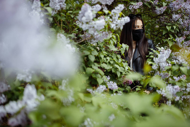 Girl in a medical mask on a background of blooming lilacs. Black mask. Protection against virus, flu. Coronavirus protection. epidemic of coronavirus. the aroma a tree in the garden Spring allergy. - Foto, Imagen