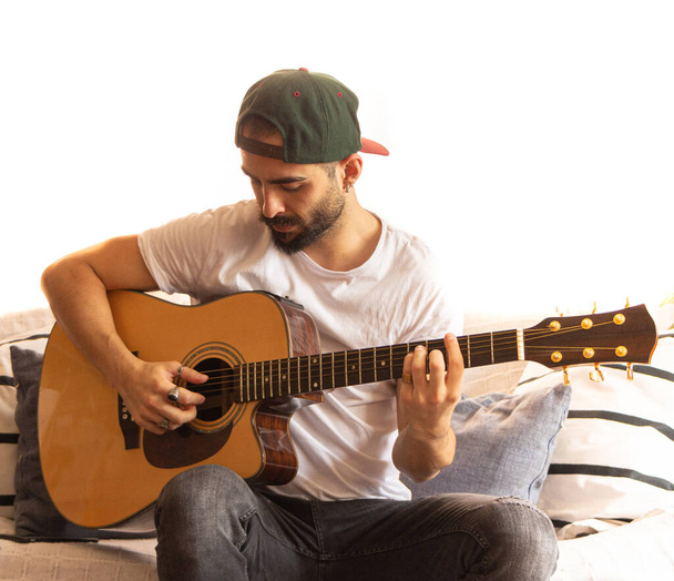 キャップと白いTシャツを着た若い男がソファの上でギターを演奏することに焦点を当てた。白の露出過多の背景。選択的焦点. - 写真・画像