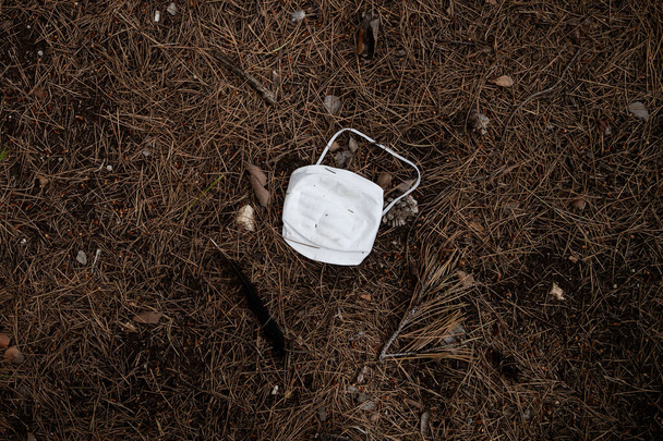 Μια μάσκα προσώπου που έμεινε στο έδαφος σε ένα πευκοδάσος. Οι μάσκες, τα γάντια και οι φιάλες απολυμαντικού που χρησιμοποιούνται κατά της εξάπλωσης του COVID-19 ρυπαίνουν το περιβάλλον. - Φωτογραφία, εικόνα