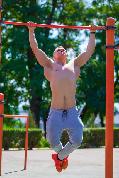 Ισχυρός μυώδης άντρας με γυμνό κορμό τραβά τον εαυτό του σε μια εγκάρσια δοκό. Γυμνάζεται σε εξωτερικούς χώρους στο καλοκαιρινό πάρκο - Φωτογραφία, εικόνα