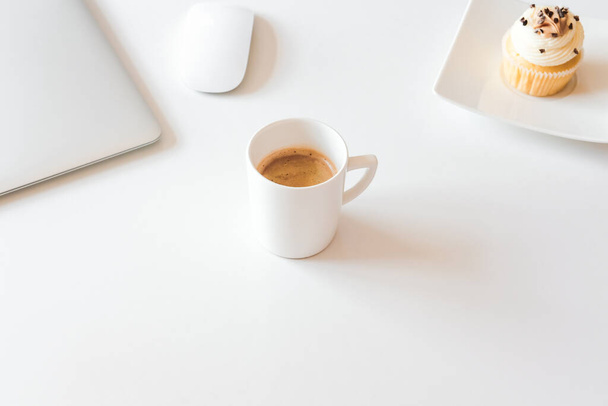 Kopje koffie en cupcake, zilveren laptop op wit bureau. Koffiepauze in huis kantoor concept.  - Foto, afbeelding
