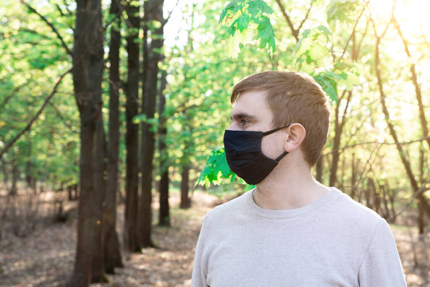 Een jongeman met een zwart medisch masker in een natuurpark. bescherming tegen quarantaine-infectie. close-up portret - Foto, afbeelding