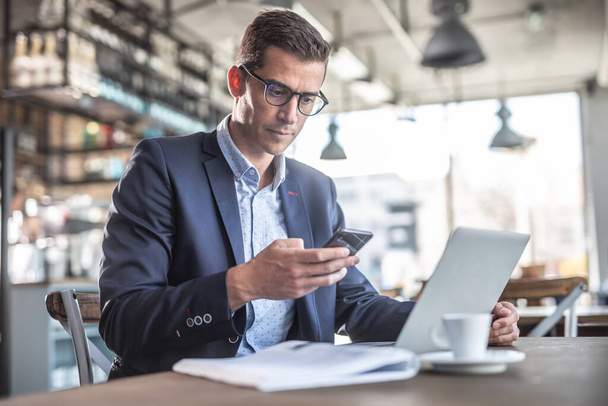 Empresario trabajando en un centro de coworking sostiene un teléfono celular con un portátil, bloc de notas y un café frente a él
. - Foto, Imagen