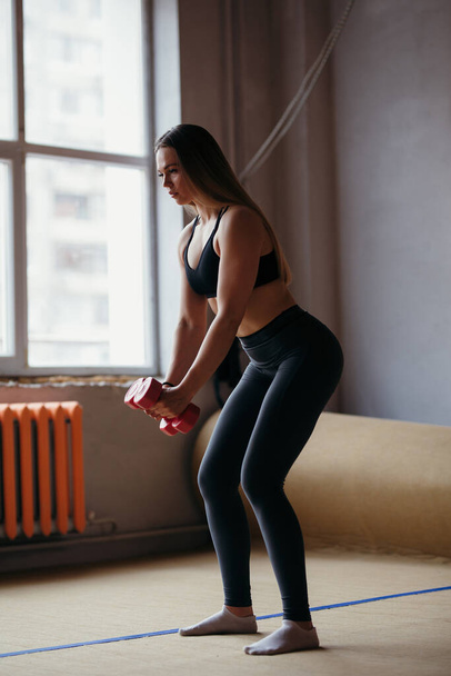 An athlete is standing next to windows of the house in quarantine squatting with dumbbells in her hands and strengthen her muscles - Fotoğraf, Görsel
