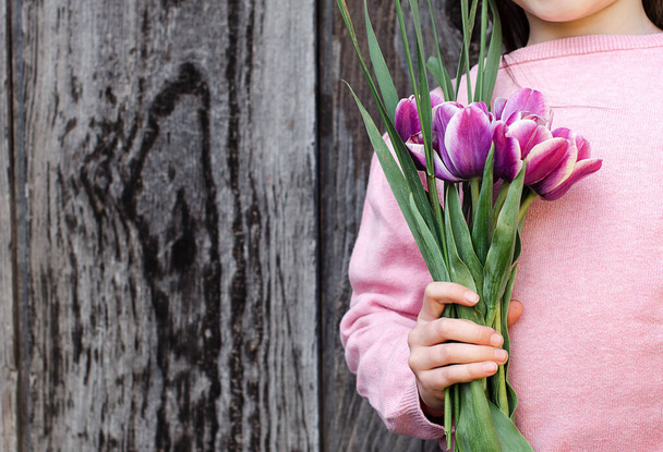 Niña con el pelo largo en un suéter rosa sostiene un ramo de tulipanes morados. Vista frontal. Antecedentes madera vieja.Concepto pring. Copiar espacio
. - Foto, imagen