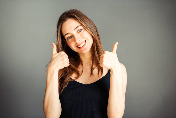 Meisje in een zwart topje met haar haar duim omhoog. Happy Op een grijze achtergrond. Zonder make-up, zonder retoucheren. - Foto, afbeelding