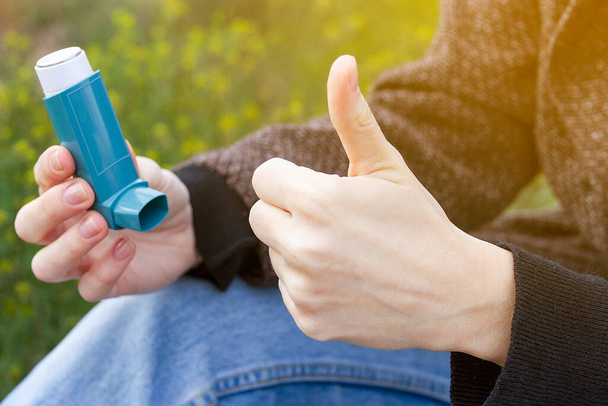 Девушка показывает большой палец вверх, держа ингалятор от астмы на цветущем фоне
. - Фото, изображение