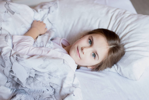 Nettes kleines Mädchen im Bett wachte am Morgen auf. Kind legt sich auf ein Kissen und blickt in die Kamera. Quarantäne-Zeit. Komfort zu Hause. Zeit zu Hause verbracht. - Foto, Bild