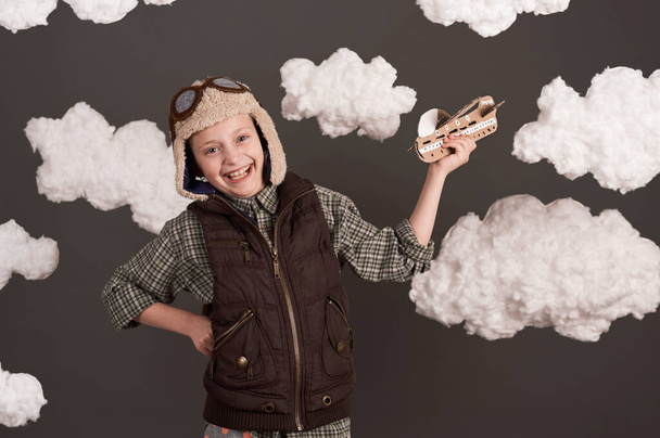 egy lány játszik egy karton repülőgép és az álmok, hogy lesz egy pilóta, öltözött retro stílusú kabát és sisak szemüveggel, felhők pamut gyapjú, szürke háttér, barna színű - Fotó, kép