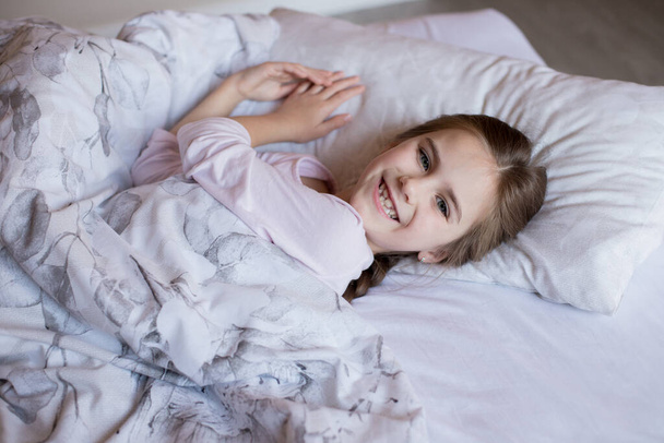 Cute little girl in bed woke up in the morning and smiles. Child lies on a pillow and looks at the camera. Quarantine Time. Comfort in home. Time spent at home. - Фото, зображення