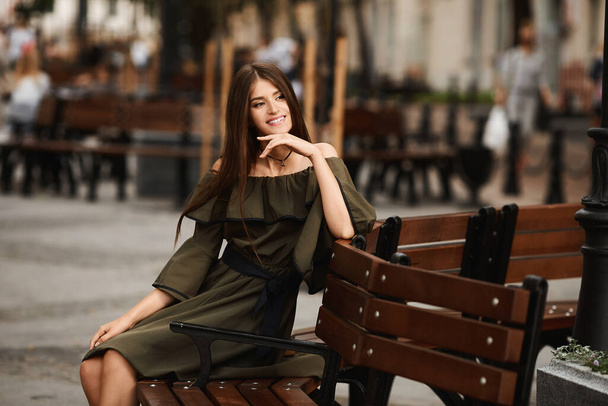 Model girl in a dress is sitting on the brown bench in the city on a sunny summer day. Outdoors lifestyle fashion portrait. - Foto, Imagem