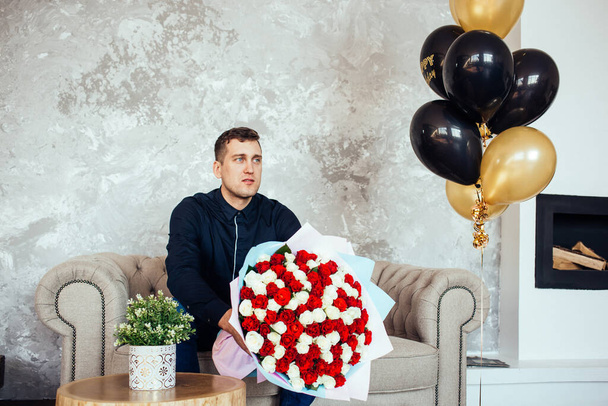 Sinipaitainen mies, jolla on iso kimppu punaisia ja valkoisia ruusuja, istuu sohvalla kotona. Hyvää syntymäpäivää. Musta ja kulta syntymäpäivä ilmapalloja. Mukavuutta kotona. Odotan vaimoa. Elämäntapa
.  - Valokuva, kuva