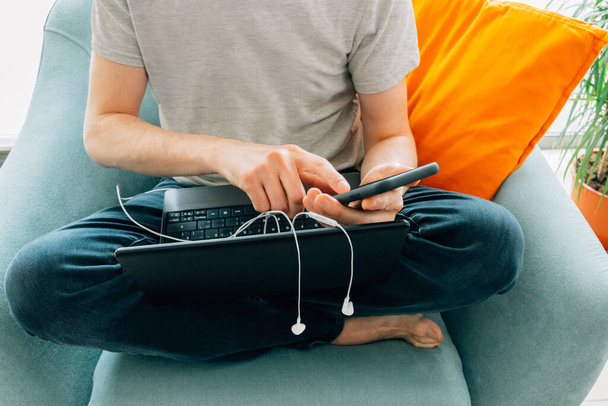 Человек, печатающий на смартфоне и ноутбуке, с наушниками и босиком на бирюзовом диване с оранжевой подушкой
 - Фото, изображение