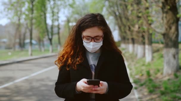 Egy fiatal lány orvosi maszkban okostelefont használ a parkban. 4k - Felvétel, videó
