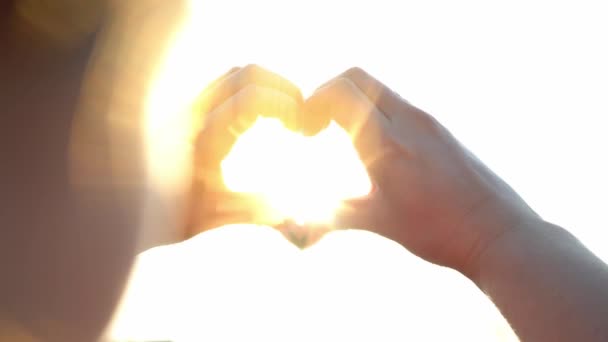 Vrouw maakt handen in de vorm van een hart in de felle zon. De magische stralen van de zon schijnen door je vingers. Fantastisch fel licht. Symbool van de liefde. close-up van aantrekkelijke vrouw handen. - Video