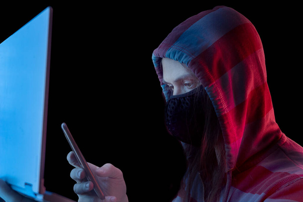 A male hacker in a sweatshirt with a hood sits in a room behind a laptop holding a phone in his hand. Cyberpunk style - Photo, Image