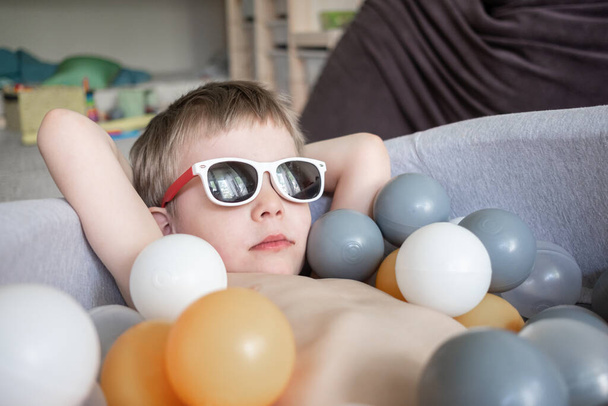 toddler boy in sunglasses swimming in ball pool at home. Self isolation quarantine entertainment at home. Holiday tourism in home - Fotografie, Obrázek