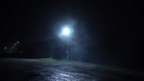 Sisli bir akşamda karanlık bir sokağı aydınlatan bir sokak lambası. Gece sisli bir sokakta. Tek direkli sisli sonbahar yolu. Çöl Sokağı. - Video, Çekim