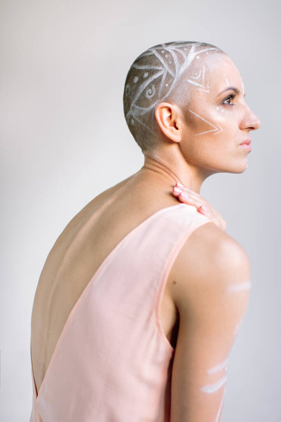 Seitenansicht einer Frau mit gemaltem Ornament auf rasiertem Kopf, die die Schulter berührt und vor grauem Hintergrund wegsieht - Foto, Bild