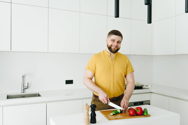 Красивий білий чоловік у жовтому різанні овочів для їжі або салату в сучасній білій кухні посміхається
 - Фото, зображення