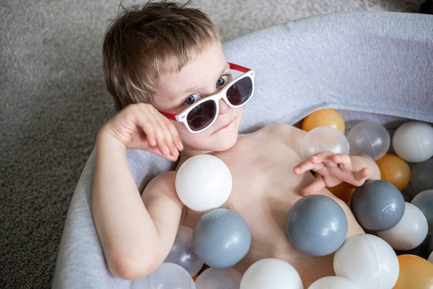 toddler boy in sunglasses swimming in ball pool at home. Self isolation quarantine entertainment at home. Holiday tourism in home - Photo, Image