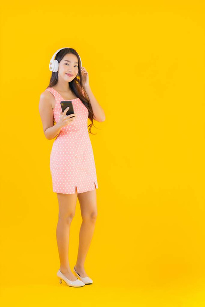 Πορτρέτο όμορφη νεαρή Ασιάτισσα γυναίκα ακούστε μουσική με έξυπνο κινητό τηλέφωνο και ακουστικά σε κίτρινο απομονωμένο φόντο - Φωτογραφία, εικόνα