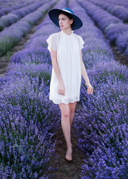Portrait of a beautiful woman in full height. The young slim model stands barefoot in a white dress and a blue hat in the middle of the beds with blossoming lavender stretching to the horizon. - Valokuva, kuva