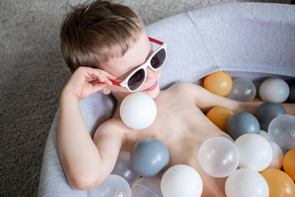 toddler boy in sunglasses swimming in ball pool at home. Self isolation quarantine entertainment at home. Holiday tourism in home - Valokuva, kuva