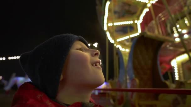 Egy fiú nézi a megvilágított körhintát esténként. Gyermek az örvénylő körhintához közel egy vidámparkban. Gyerekek karácsonyi körhinta koszorúban. Szórakoztató családi nyaralás. Boldog gyermekkort!. - Felvétel, videó