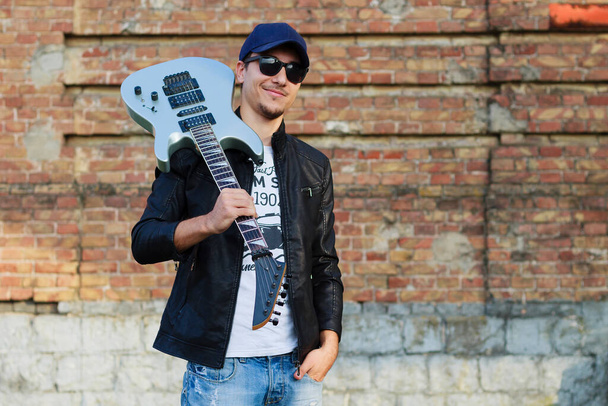 Νέος όμορφος άντρας με μια κιθάρα στον ώμο του, το θολό φόντο ενός τοίχου από τούβλα - Φωτογραφία, εικόνα