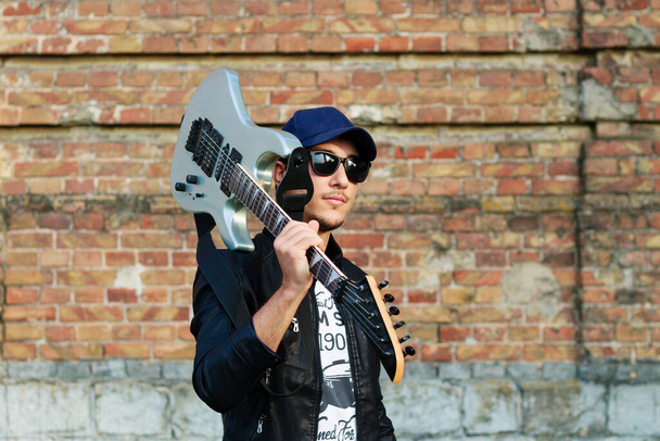 Mladý pohledný chlapík s kytarou na rameni, rozmazané pozadí cihlové zdi - Fotografie, Obrázek